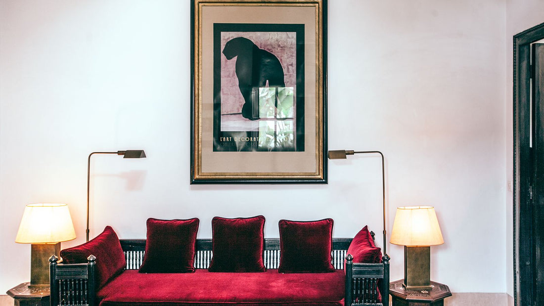 A burgundy velvet sofa with red cushions beneath a painting on a cream wall, framed by two lamps