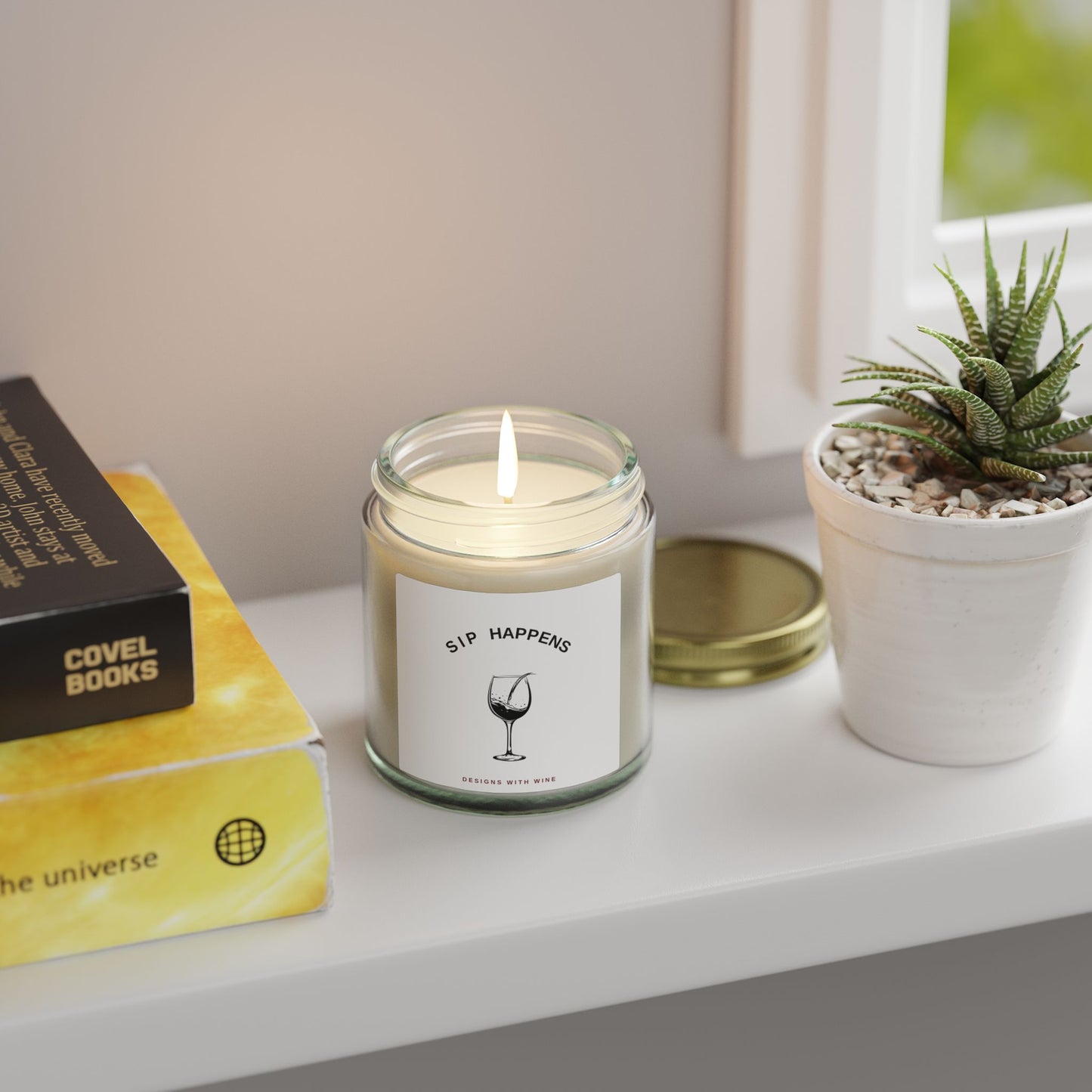 Sip Happens candle lit on a windowsill beside stacked books and a potted succulent